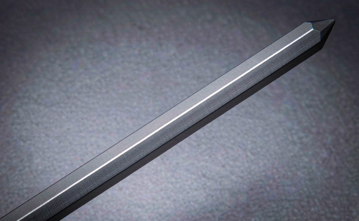 A faceted metal rod lying diagonally on grey paper, flat faces and edges clearly visible